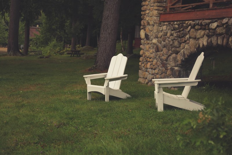 two white lawn chairs beside a stone house next to a forest and picnic chairs, lawn chairs 4k
