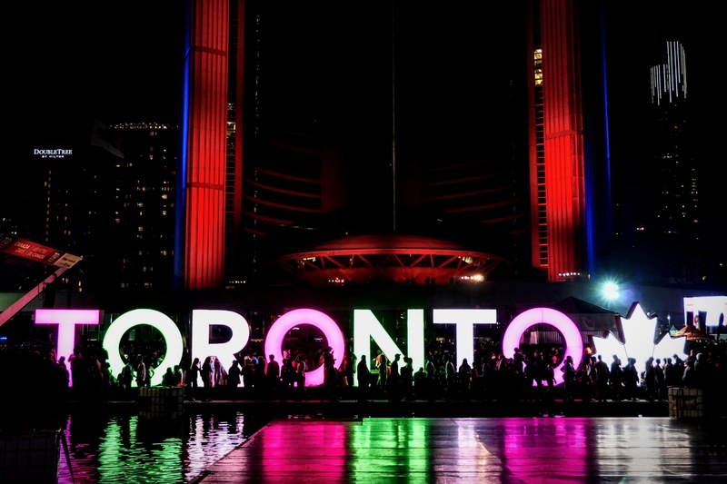 crowd of people gathers by a neon sign that says toronto at night, reflecting neon lights 4k