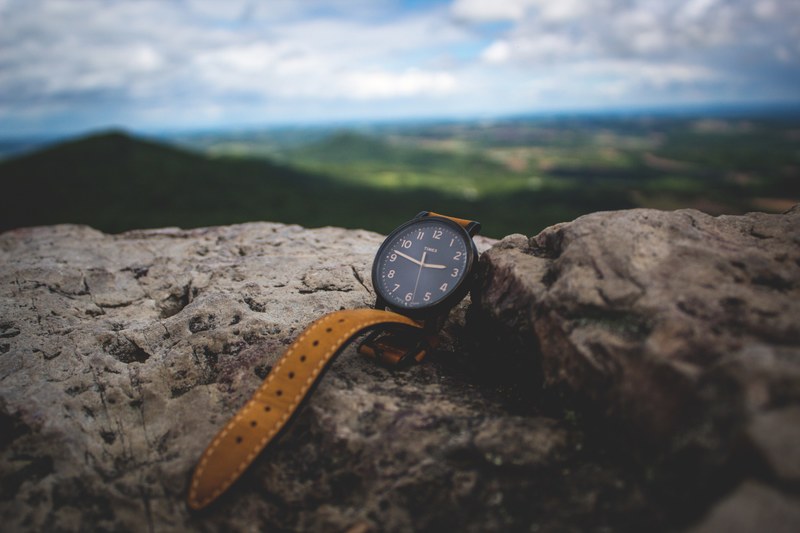 an analog watch with a brown leather strap and a black face sitting on a stone overlooking green mountains and a cloudy blue sky, watch in the mountains 4k