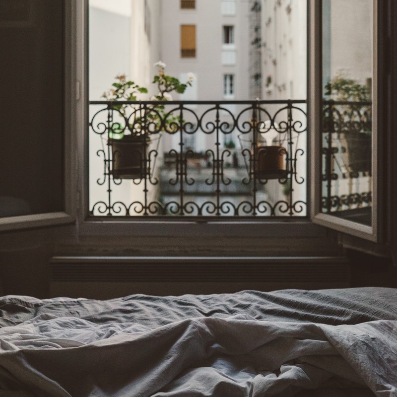 an open window at the foot of an bed with messy sheets looks at flowers on the sill and other buildings across the way, open bedroom window 4k