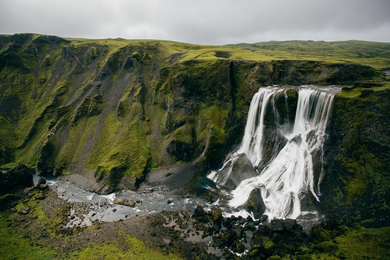 waterfall rushing down a green mossy cliff with a grey cloudy sky in the background, mossy rock mountain and waterfall 4k