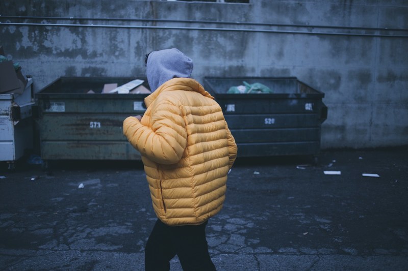 a person in a yellow puffy jacket and hood walks in an alley next to dumpsters, person in alley with dumpsters 4k