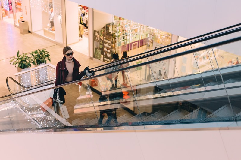 chic woman in sunglasses holds bags up a mall escalator, mall shopping 4k