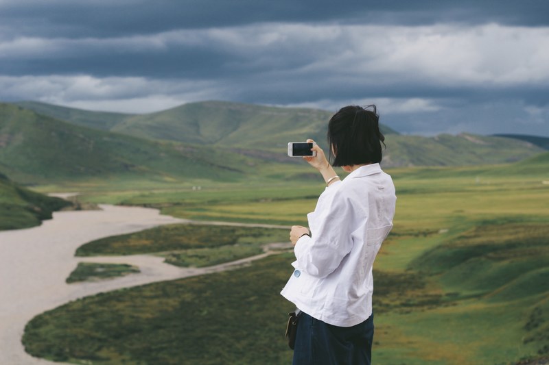 person in white shirt using a smartphone to photograph green hills and river, taking photo of hills 4k