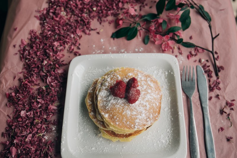 delicious looking pancakes covered in berries and powdered sugar on a plate that is sitting on a table with flowers on it, millennial pink pancakes 4k