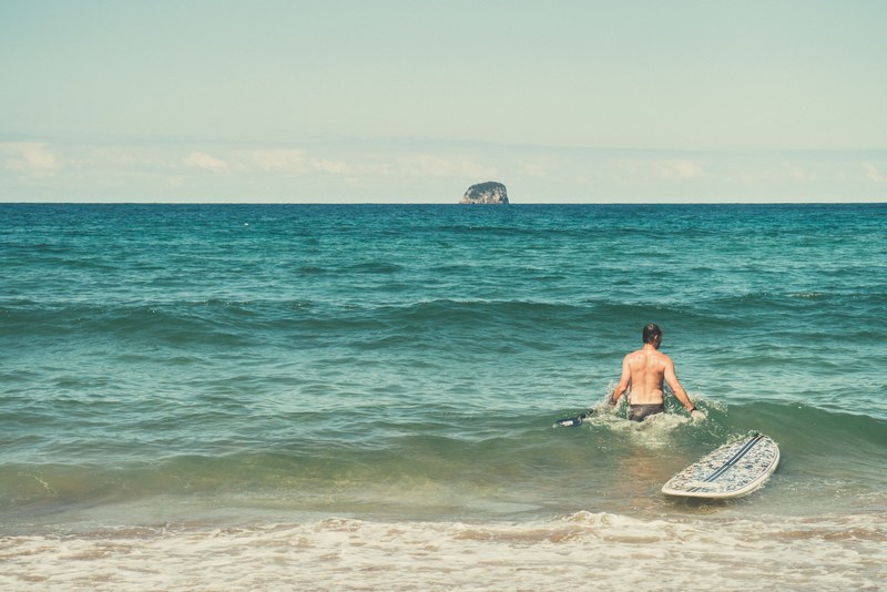 surfer with a surfboard entering the calm ocean, surfer looking at the rock formation 4k