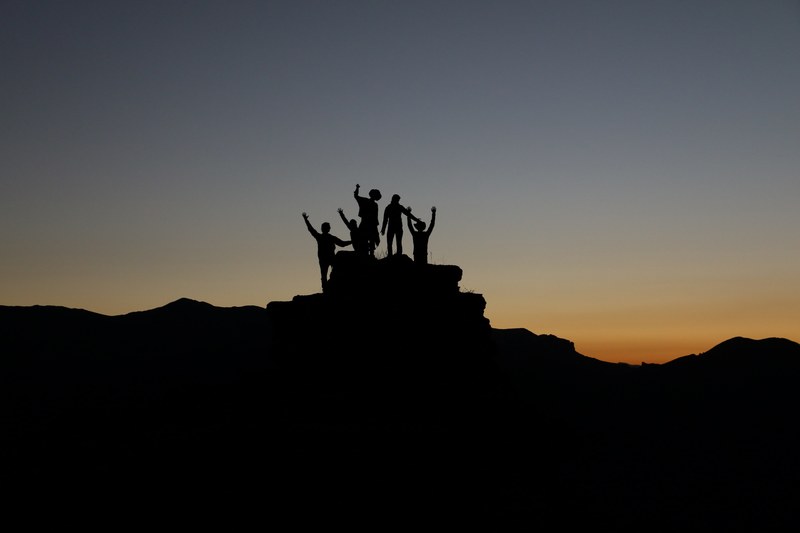silhouette of a group of friends with arms raised atop a mountain peak during sunset, we did it 4k