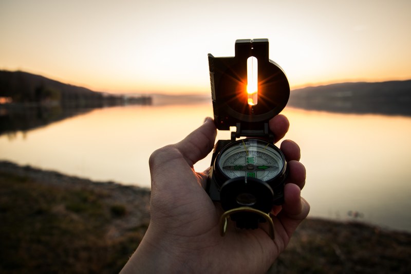 a hand holding up a lensatic compass and pointing the lens at the setting sun, setting sun in the west 4k