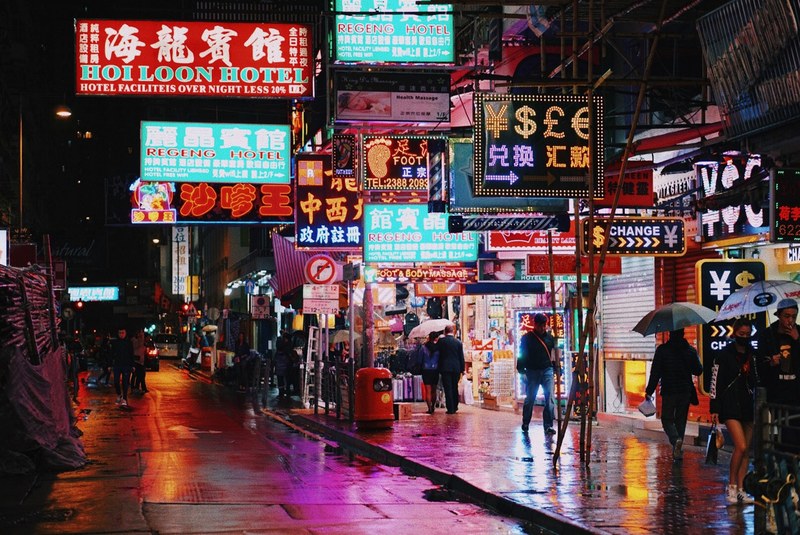 night time scene of a city street with several neon lights and signs over people walking around with umbrellas in a hong kong urban region, hong kong neons 4k