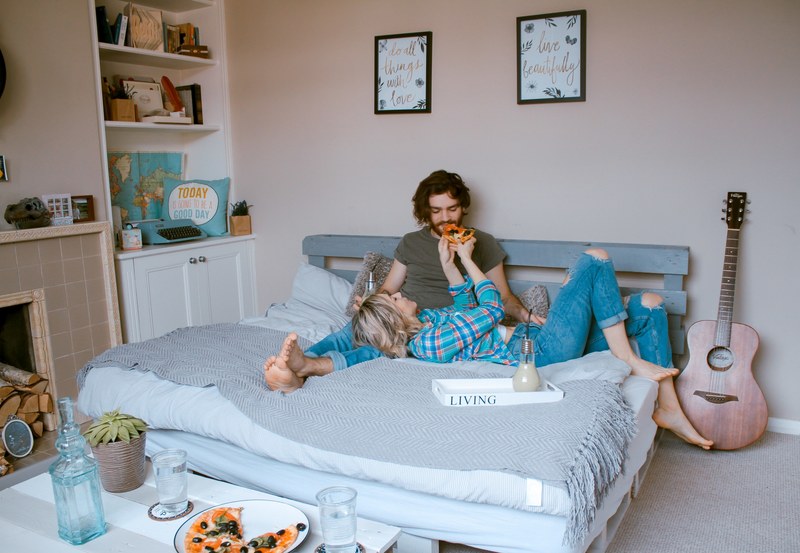 young couple on bed feeding each other pizza with fireplace guitar and wall art in teddington, pizza always in the way 4k