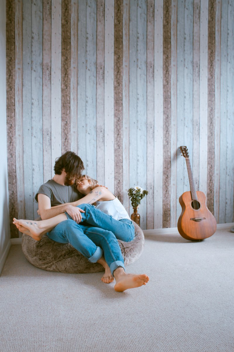 a barefooted man and woman cuddle on a beanbag chair against wood paneling near a guitar, still in your arms 4k