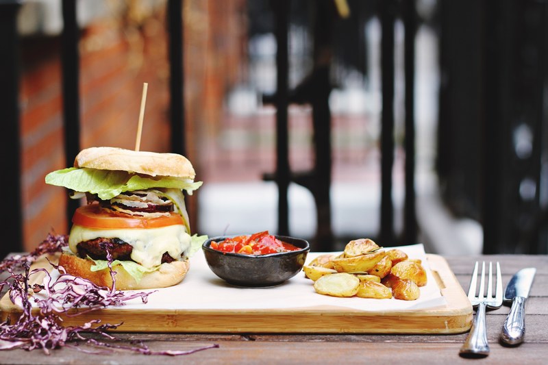 a chopping board with a handmade cheeseburger salsa and sliced potatoes beside a fork and knife on a wooden table, giant burger 4k
