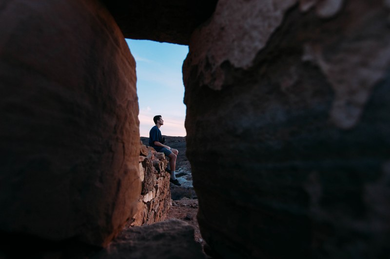 a man sitting on red rocks seen through a gap in a rock formation, man seated on red rocks 4k