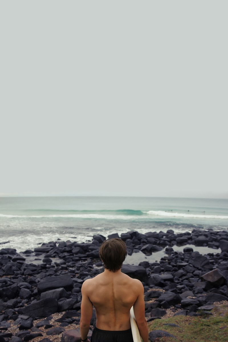 topless man with a surfboard on a rocky coast of burleigh beach, over the rocks and into the sea 4k