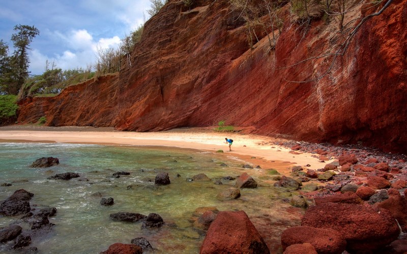 a person standing on a rocky beach under a steep red rock face, rocky beach under a red cliff 4k