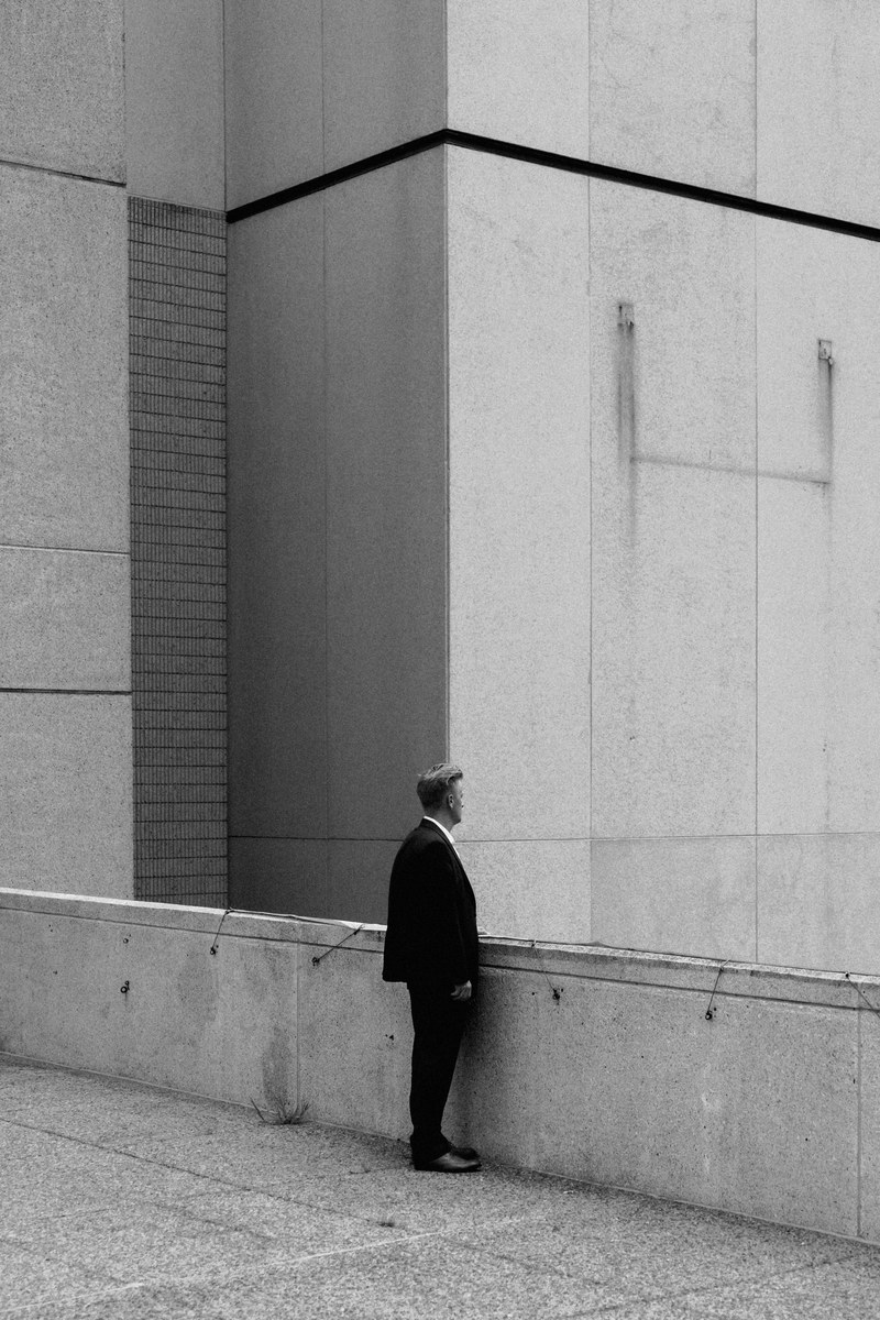 a black and white shot of a man in a black suit standing near a building ledge, lone man amongst concrete 4k