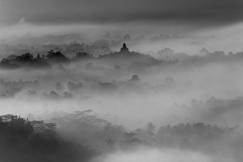 a desaturated shot of the silhouettes of wooded hills with a spire of a temple rising up above the mist, borobudur in misty morning 4k