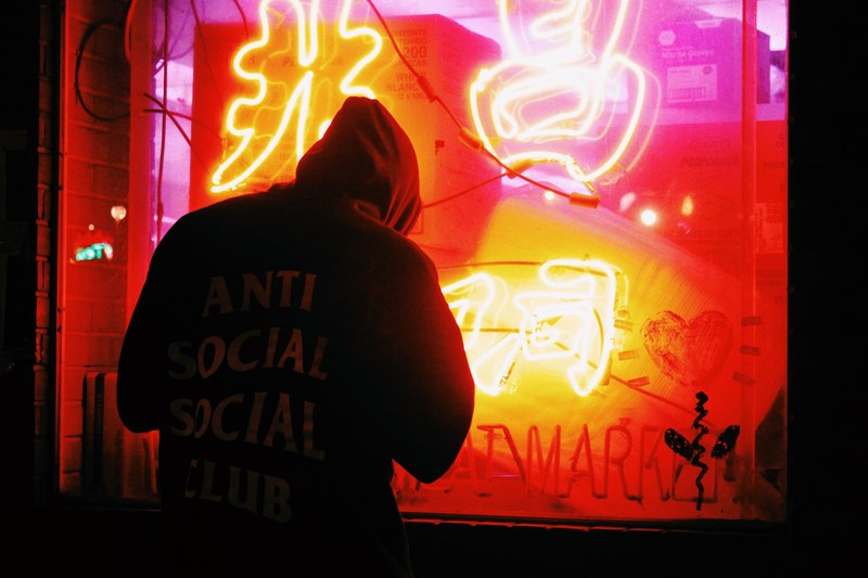 man in hoody with anti social slogan standing outside red neon display in store window, man red neon window display 4k