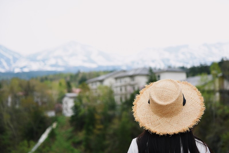 a woman with dark hair wearing a straw hat looking at a village surrounded by trees under a snowy white mountain, girl 4k
