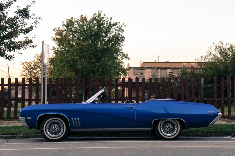 blue convertible vehicle parked on pavement in front of brown picket fence trees and building, blue convertible brown fence 4k