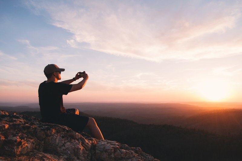 a man sitting on jagged rocks photographing the wooded hills below at sunset, photographing the sunset 4k