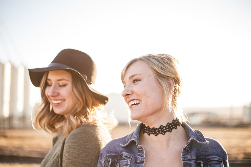 two women in a hat and choker stand closely laughing against a blurry outdoor background, women with hat and choker 4k