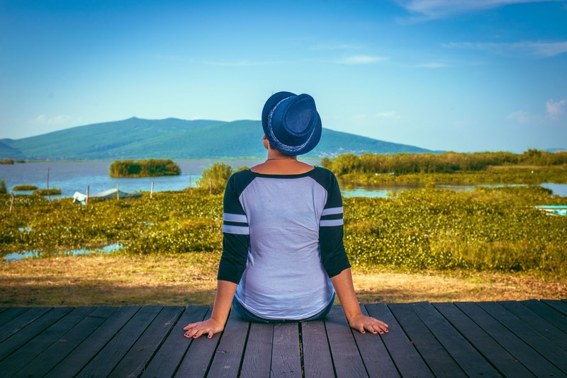 a person sits on the edge of a boardwalk looking out at a marshy landscape, looking out at marsh 4k