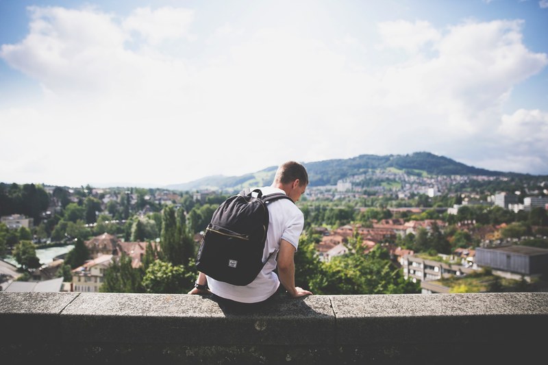 a man with a backpack sitting on a ledge overlooking a picturesque town, vacation in a pleasant town 4k