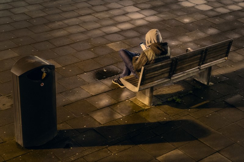 a hooded man on a bench using his smart phone beside a garbage can in a tile floor outside at night, hooded man on bench 4k