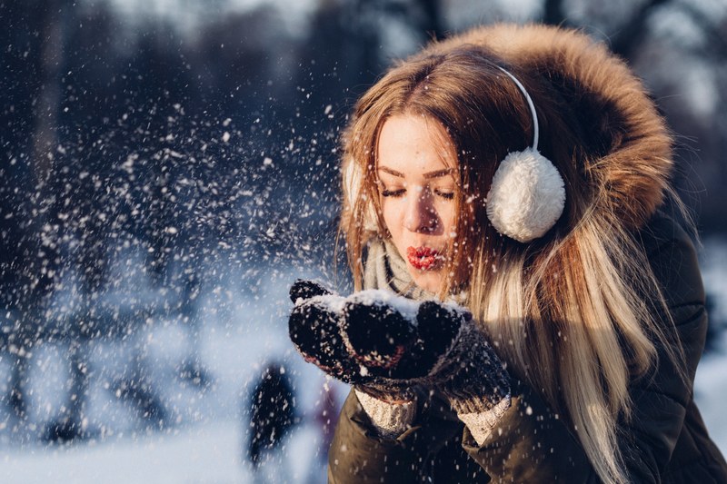 a woman wearing earmuffs a winter coat and red lipstick blowing snow out of her hands in olsztyn, snow in mittens 4k