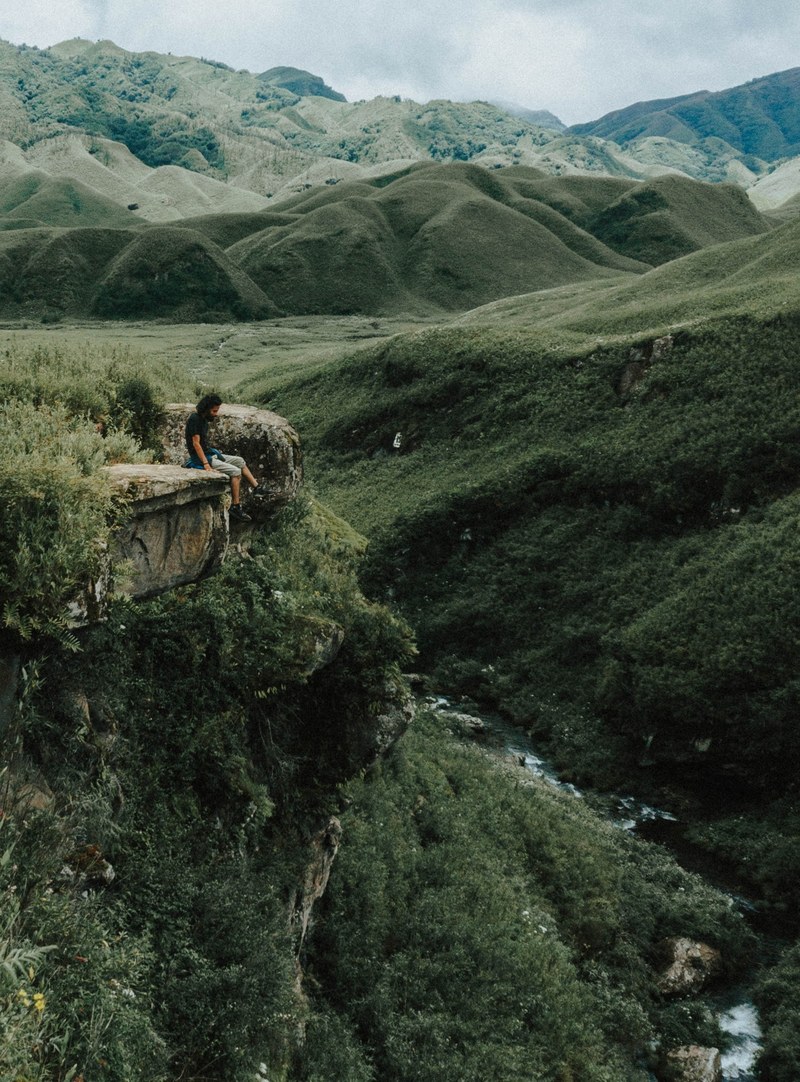 man sits on the rocky ledge of a mossy hillside by a creek, wilderness 4k