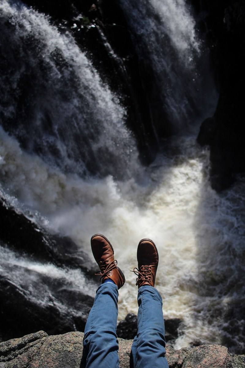 a males legs in blue pants and boots hanging over the side of a waterfall, shoes and waterfall 4k