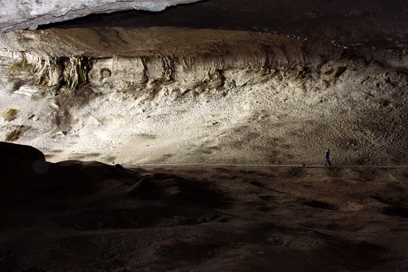 a person walking on a path cutting through a large cave with sun coming through the entrance, exploring the cave 4k