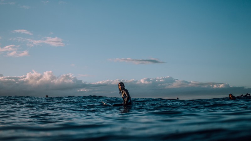 a female surfer sat on her board in the calm water and under a blue cloudy sky at piha with other surfers in the background, should be a corona ad 4k