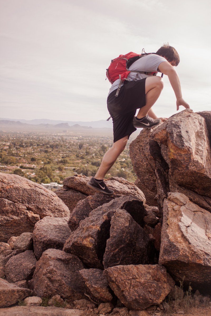 a person wearing a red backpack on a hike climbs on a pile of rocks, i can jump that far 4k