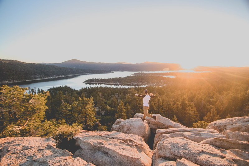 a person jumping on a rocky ledge overlooking a forest and a lake, big bear lake 4k