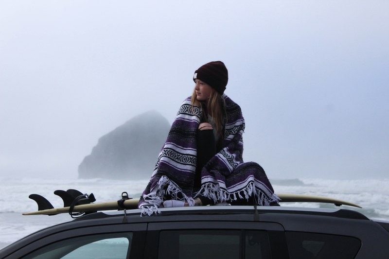 woman wrapped up in a blanket sitting on a car roof by the surfboard at cannon beach, woman sitting on a car roof 4k