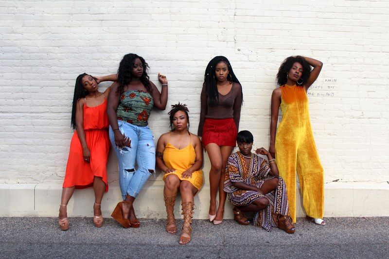 group of women standing and sitting against a white brick wall on the sidewalk in uptown, reflecting queens 4k