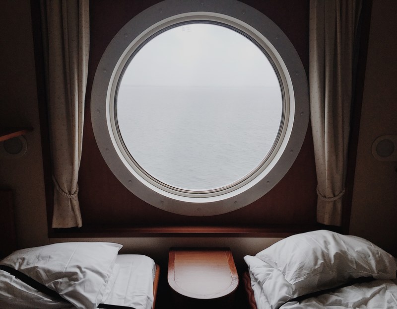 two beds with pillows on either side of a circular window with curtains overlooking water, circular window and beds 4k