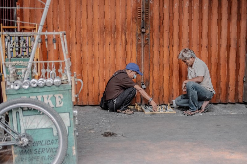 two men playing chess while sitting on the floor in front of a red building next to a bike store, chess on the run 4k