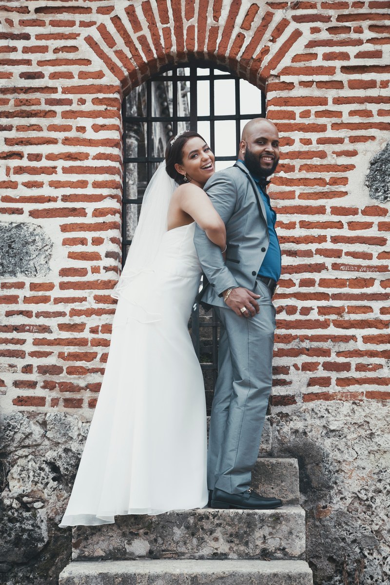 a bride holding a grooms arms from behind on concrete steps by a brick wall, bride wrestles groom 4k