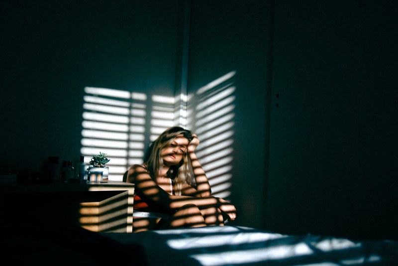 a dejected looking woman sat in a dark bedroom with her hand on her head with horizontal strips of light shining on her from a blind bloomington, we are all human 4k