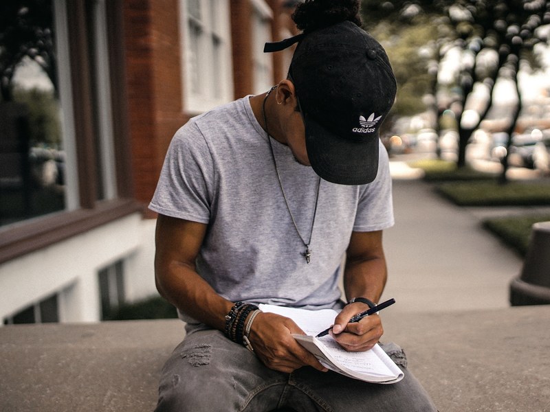 a man in a baseball cap taking notes while sitting on a concrete ledge outdoors, heart on paper 4k