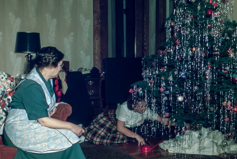 a classic photo of a mom wearing a pinafore and glasses sitting on a chair watching her daughter put gifts under a christmas tree, putting gifts under the tree 4k