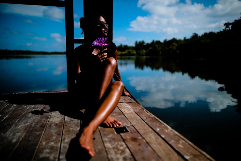 a woman holding a flower sits on a dock overlooking the water which reflects blue skies, maldives 4k