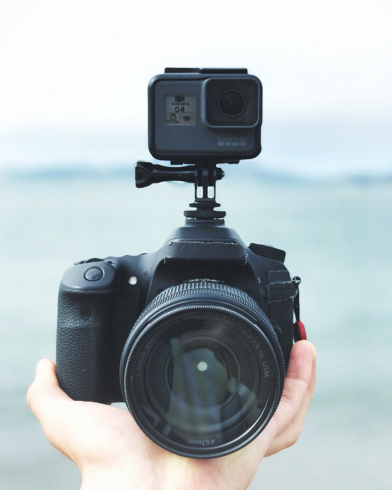 macro of a person holding a black dslr camera with a gopro mounted on beside a lake or sea setting, the rig 4k