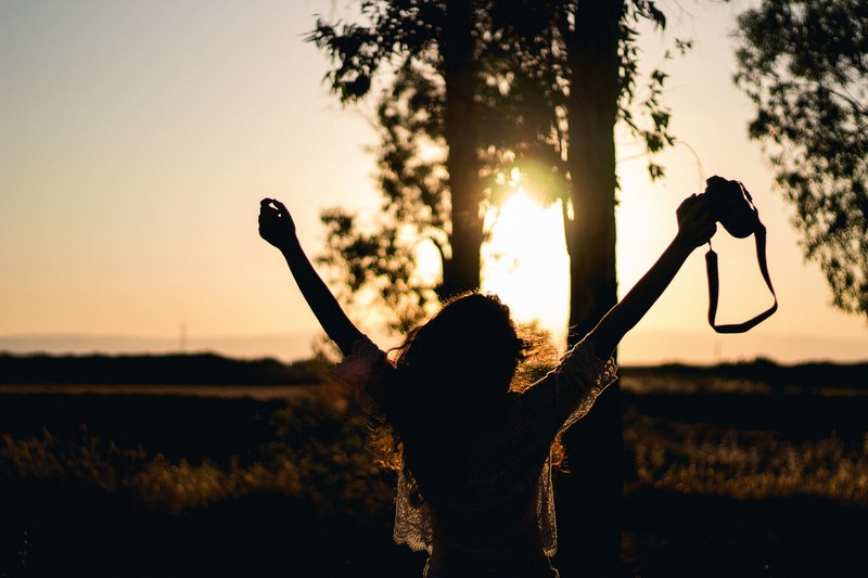 a woman stretches her arms in the air holding a camera as a golden sunset illuminates the sky, livin free 2 4k