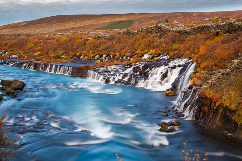 hdr photo of whitewater waterfalls from a hilly rocky edge beside a forest of autumn trees cascading down a clear and fast moving river, hraunfosar in the fall 4k