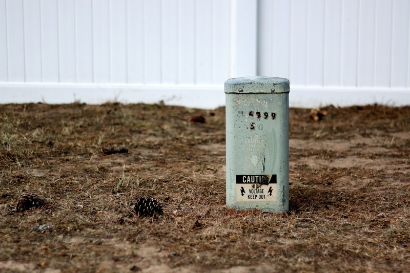 an old and rusted power box with a high voltage warning sticker sitting disconnected on a pinecone littered dry ground beside a white fence, high voltage 4k
