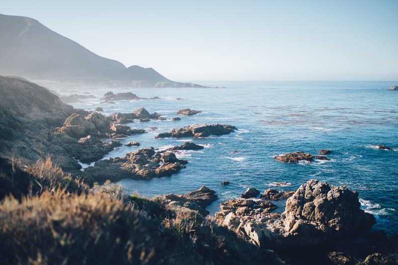 a rocky coastline with mountains in the background overlooking the blue sea at bixby creek bridge, mountains over the sea 4k